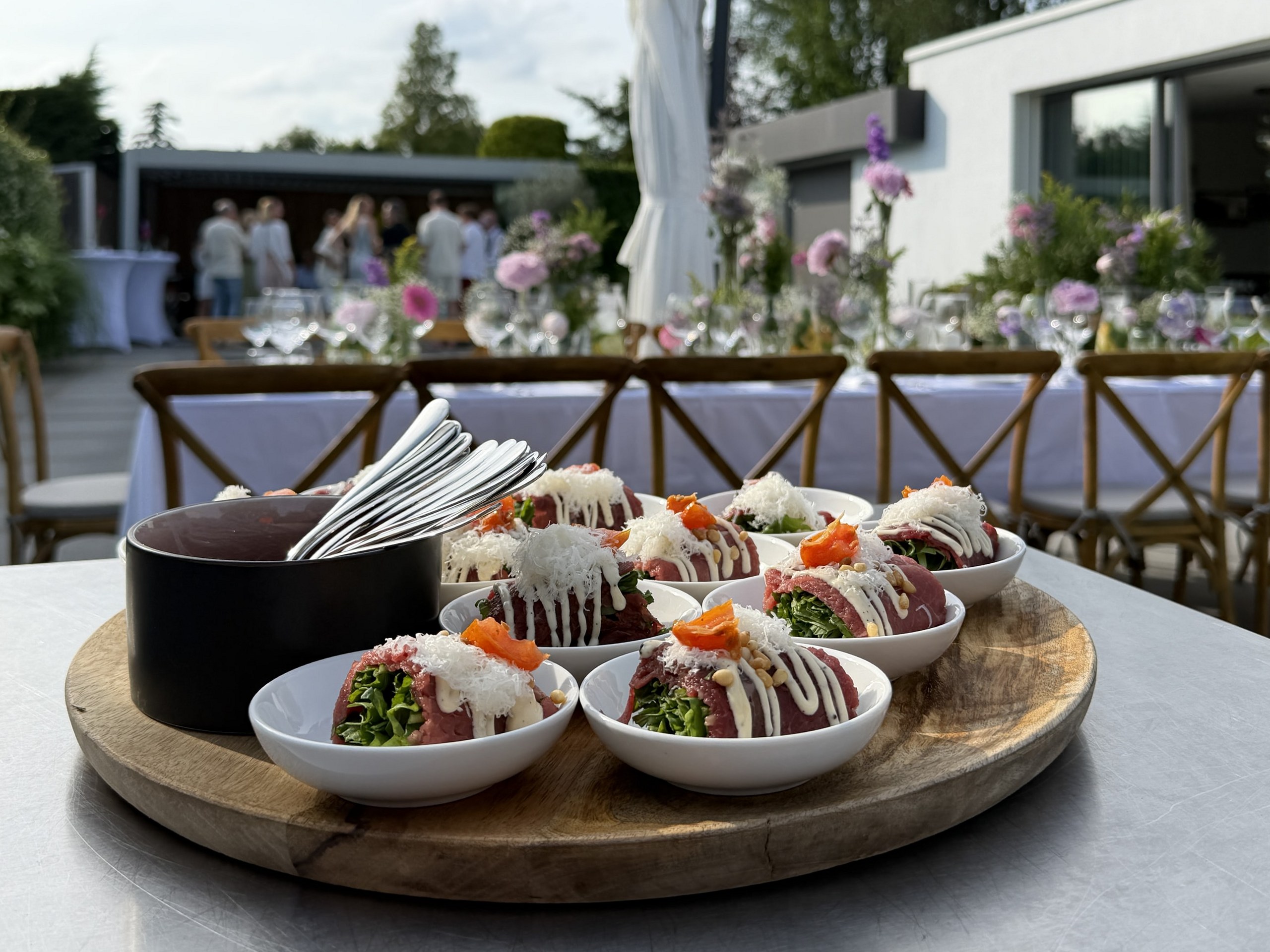 Houten plateau met kommetjes opgerolde carpaccio, buiten op een feest met gedekte tafels erachter.