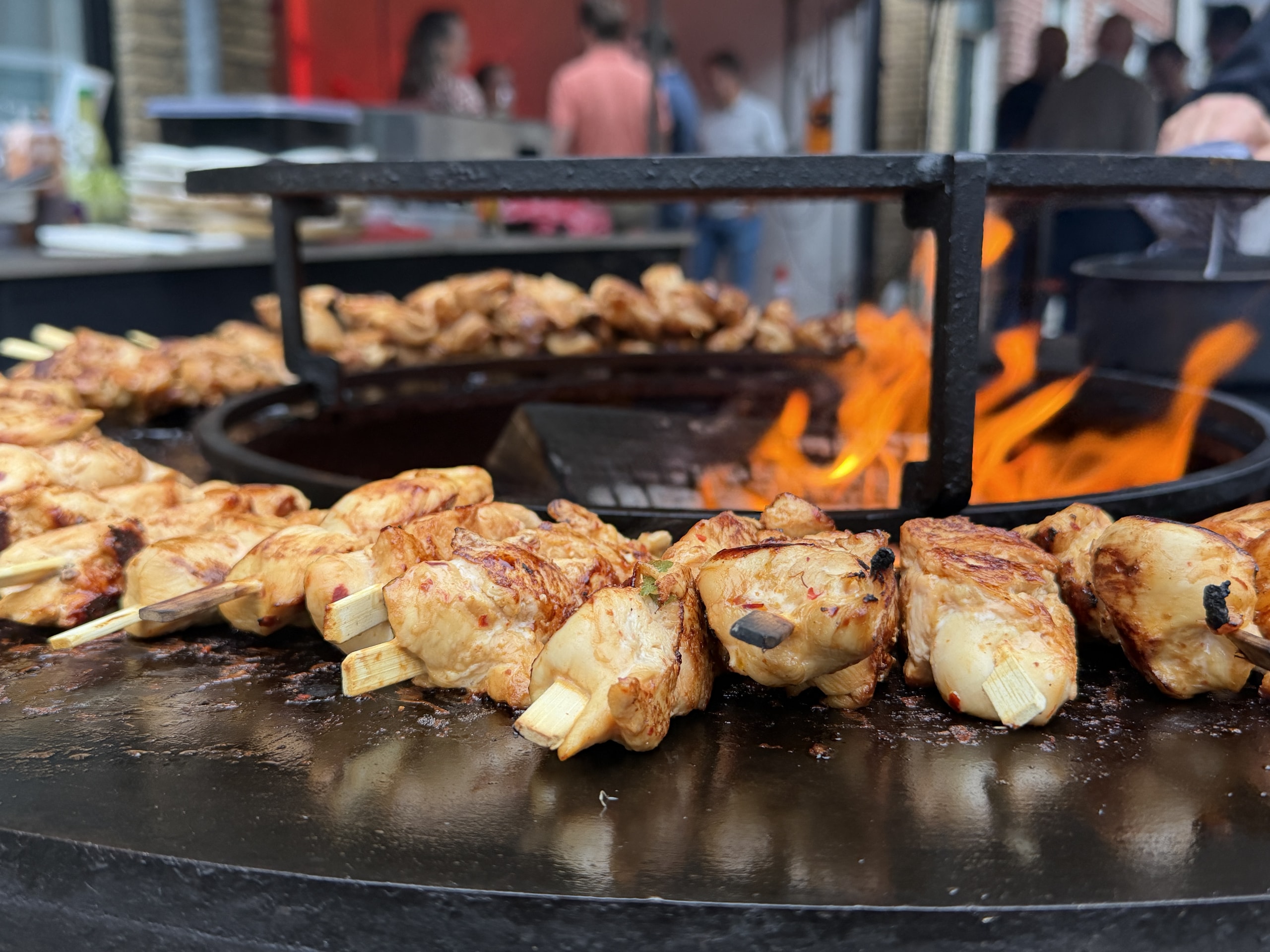 Kipspiesen op een vuurplaat met vlammen erachter, buiten op een feest met gasten op de achtergrond.