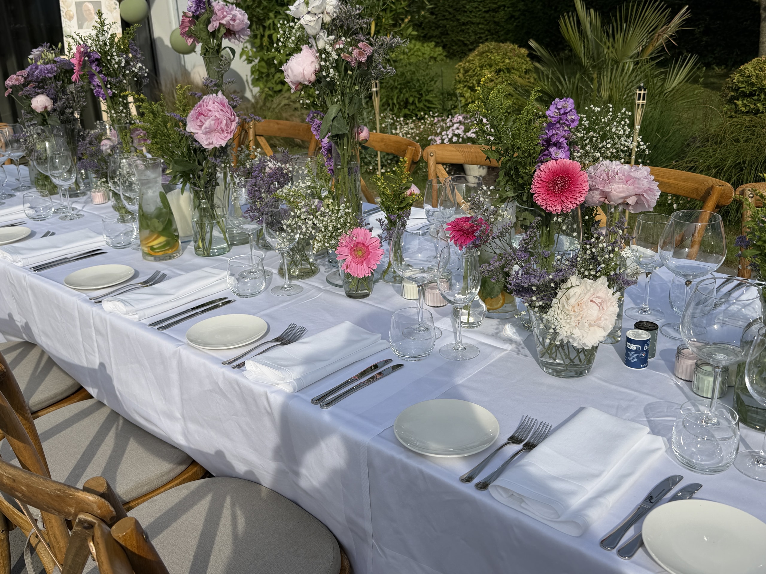 Lange gedekte tafel buiten, met glaswerk, servetten en vaasjes met veldbloemen.