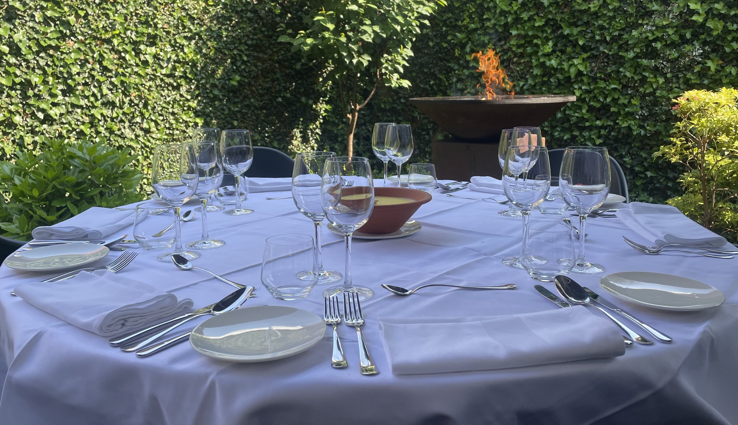 Ronde gedekte tafel met wit tafelkleed en glaswerk in een groene tuin, met een brandende vuurschaal erachter.