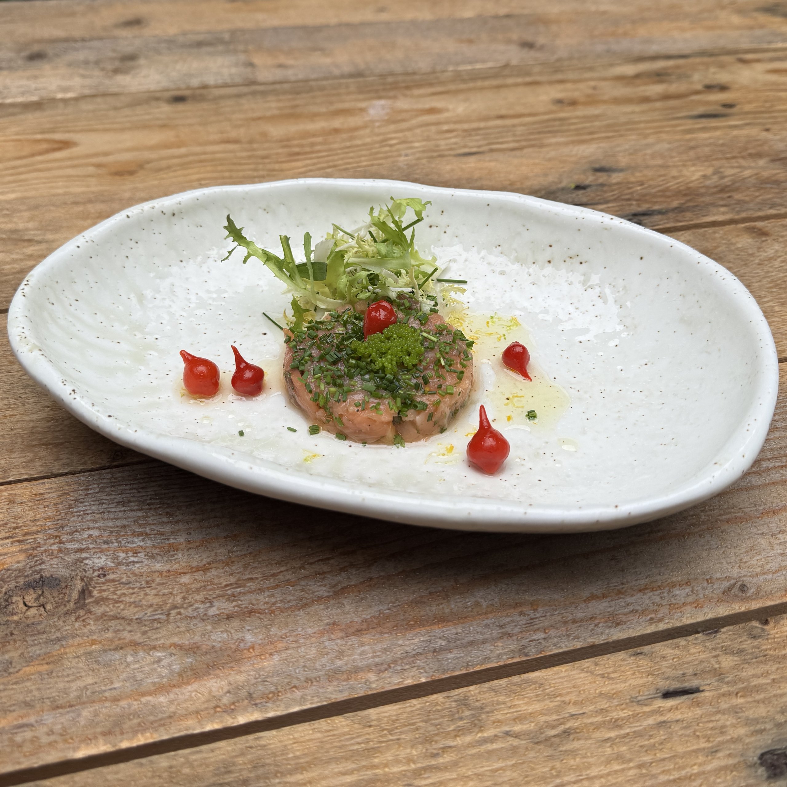 Tartaar met frisée, groene kruiden en druppels rode saus op een ovale witte schaal, op een houten tafel.