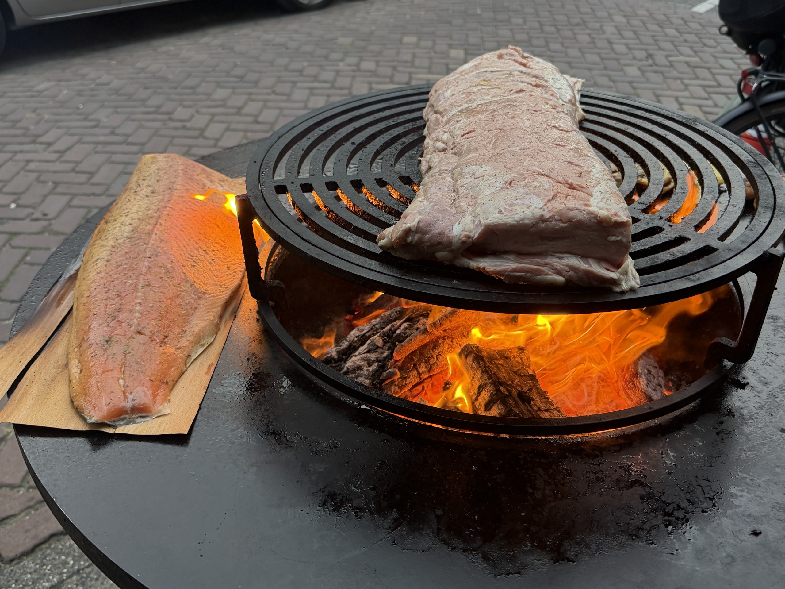 Zalmfilet op een plank en een groot stuk vlees op het rooster boven het vuur.